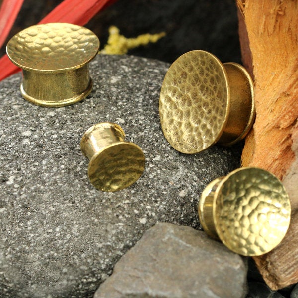 Four Hammered Round Saddle Plugs in gold, ranging from 8mm to 16mm gauge, are displayed on a textured stone surface with pieces of wood and foliage in the background. These Kaya Movement brass plugs showcase a rustic, handcrafted appearance.